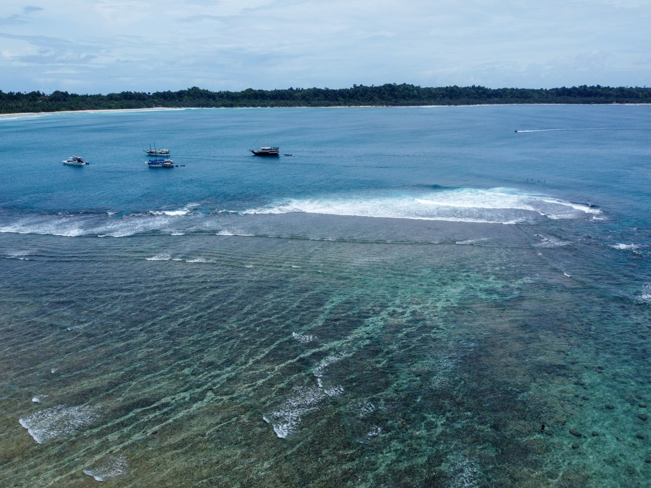 Boats anchored near coral reef with waves and distant shoreline under a cloudy sky.